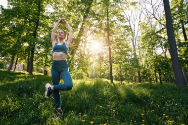 Rahatlamış genç dişi yeşil ahşap üzerinde egzersiz yapıyor ve Asana 'da elleri havada tek ayak üzerinde dengede duruyor.