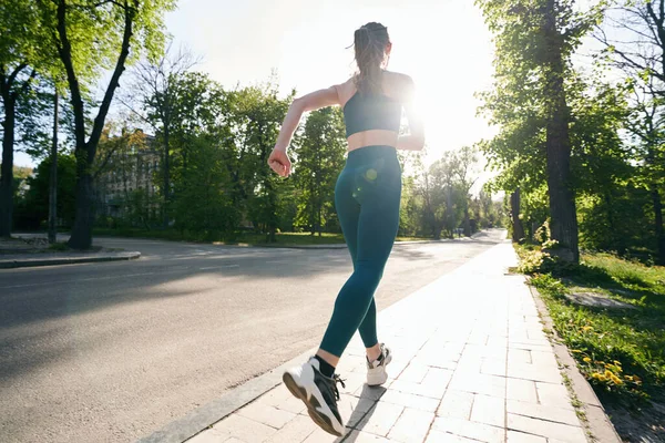 Spor giyimli atletik kadın şehir parkında güneşli bir günde antrenman yaparken koşuyor.