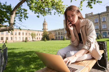 Zarif bir kadının dar açılı bir açıyla bankta oturması ve akıllı telefonda iş hakkında konuşurken not defteri kullanması.