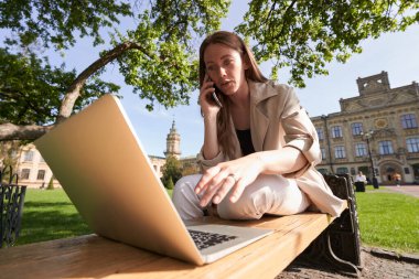 Parkta dizüstü bilgisayarla çalışan ve cep telefonuyla konuşan zarif bir kadının düşük açısı.