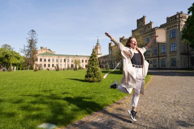 Neşeli genç bir kadın elinde defterle eski bir bina ve atlama ile parkta yürüyor.