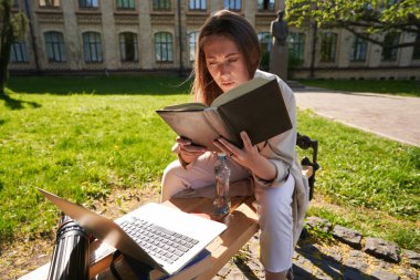 Genç bir bayan açık havada defter kullanırken elinde bir şişe su ve kitap ile bankta oturuyor.