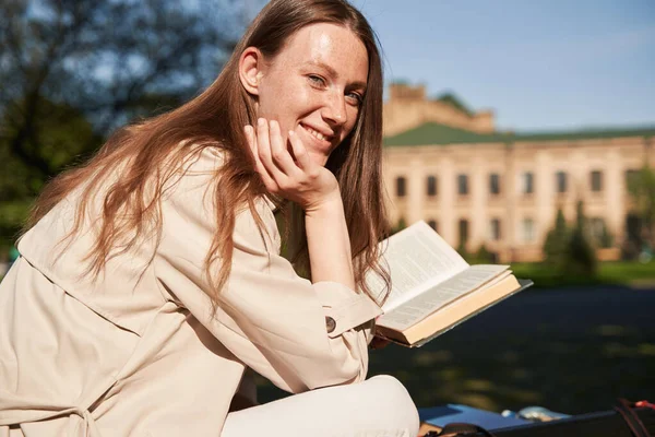 Waist up portrait of smiling lady sitting on bench with tube for draings and enjoying literature in park near university