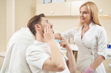 Estetik kliniğinin erkek müşterisi yüzüne işaret ediyor ve güzellik uzmanıyla konuşurken yanağına dokunuyor.