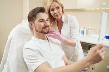 Beyaz tenli güzellik uzmanı, aynaya bakarken çenesine dokunarak erkek yüzünü düzeltiyor.