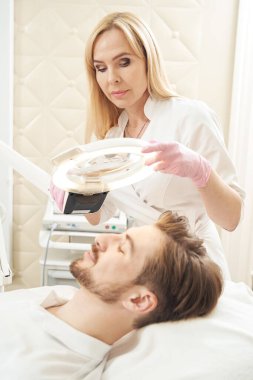 Güzellik uzmanı, estetik ameliyat sırasında erkek müşterinin yüzünün üstündeki ameliyat lambasının konumunu ayarlıyor.