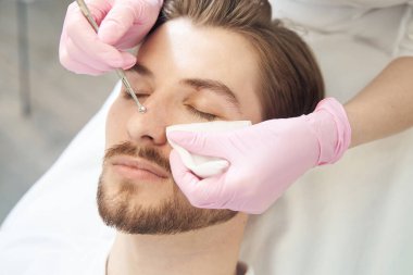Güzellik merkezindeki adam gözleri kapalı yatarken güzellik uzmanı burnunun gözeneklerini temizlemek için uno kaşık kullanıyor.