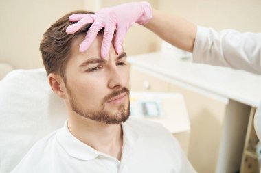 Genç bir erkek, estetik klinik çalışanı alnına şaplak atarken sandalyede oturuyor.