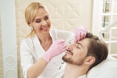 Kadın gülümseyerek erkeğin burnunun üst kısmına botulinyum toksini enjekte ediyor.