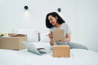Yatakta elinde karton kutuyla oturan ve dizüstü bilgisayara bakan bir kadın.