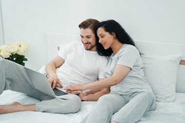 Gülümseyen mutlu kadın ve erkek yatakta oturup taşınabilir bilgisayar kullanıyor.