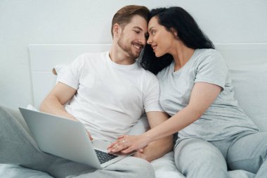 Dizüstü bilgisayarlı romantik bir genç adam yatakta mutlu bir kadının yanında oturuyor.