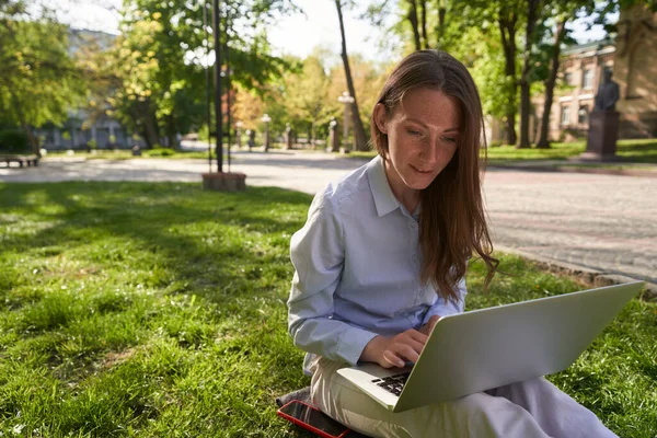 Gülümseyen kadın parkta çimlerin üzerinde oturuyor ve yanında akıllı telefonu varken defter kullanıyor.