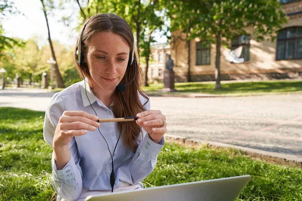 Młoda Wesoła Kobieta Siedzi Trawie Nosi Słuchawki Mikrofonem Patrząc Ekran — Zdjęcie stockowe