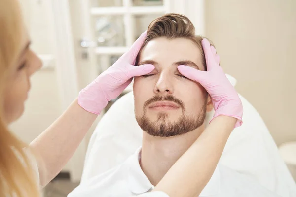 美容师用大拇指垂下男性访客的眼睑 同时研究他额头上的皮肤 — 图库照片