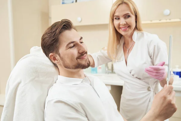 Man sitting in chair in beautician office and looking at his face in handmirror near smiling beauty therapist