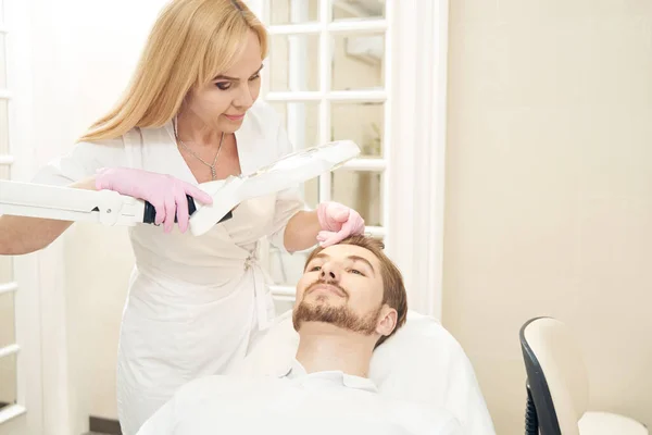 Woman beauty therapist standing over partient of aesthetic medical clinic in chair and moving hand across his forehead