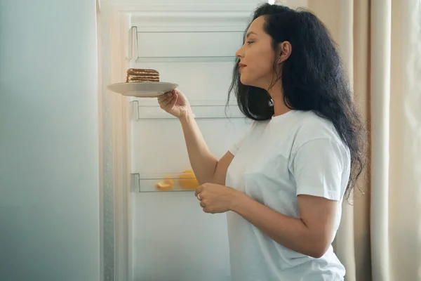 Side view of woman placing slice of chocolate cake on plate into refrigerator