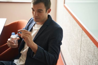 Üzgün bir erkek kâtip, elinde su şişesi tutarken parmaklarının arasından mavi hapa bakıyor.