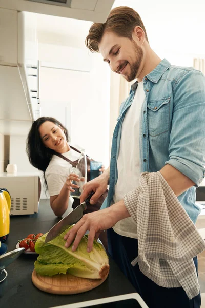 Smiling happy young male cutting cabbage on kitchen counter in presence of his life partner