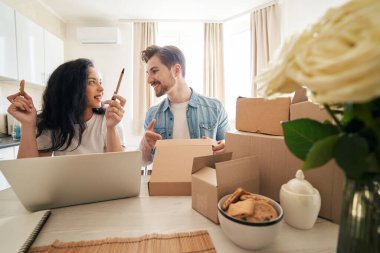 Masada oturan kadın, erkek arkadaşına bakıyor ve sipariş edilmiş eşyalar için karton kutu hazırlıyor.