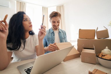 Düşünceli kadın dizüstü bilgisayar başında oturmuş, meslektaşı sipariş edilmiş eşyaları paketlemek için karton kutu hazırlarken yukarı bakıyor.