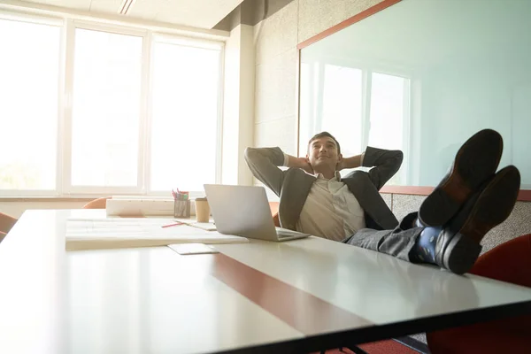 Male clerical worker leaning back in chair with hands behind head and feet on table while daydreaming