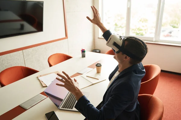 Side view of office worker raising hands and touching air while sitting at desk in virtual reality goggles