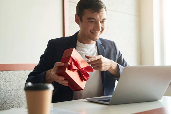 Excited male office worker showing red box with present to person online through laptop
