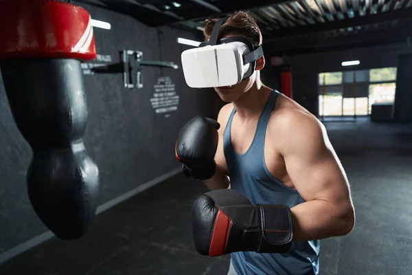 Muscular man in VR glasses and boxing gloves working out at gym