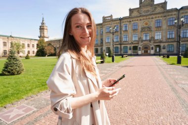 Neşeli bir kadın elinde akıllı bir telefonla çimlerle çevrili geniş bir sokakta duruyor.