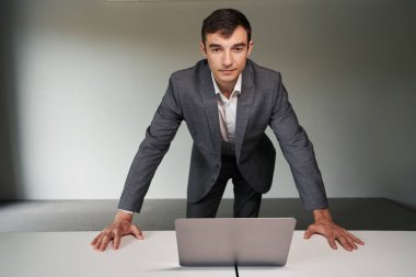 Adam ofis çalışanı dizüstü bilgisayarın önünde duruyor, elleri masanın üstünde ve kameraya bakıyor.