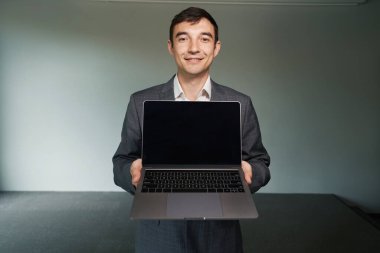 Adam ofis çalışanı yüzünde gülümsemeyle kameraya dizüstü bilgisayar gösteriyor.