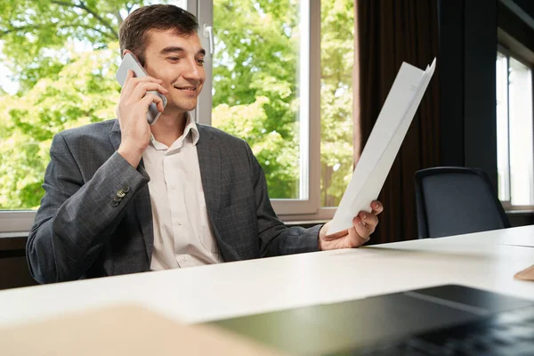 Businessman sitting at table in office room and speaking on mobile phone with official paper in hand