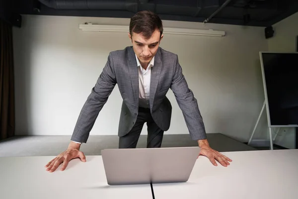 Entrepreneur in gray suit leaning upon the table with both hands while staring intensely at laptop screen