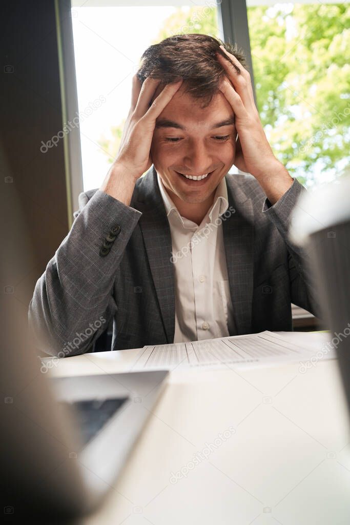 Caucasian man grabbing head and laughing hysterically while looking at spreadsheet on table before him