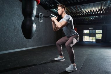 Spor salonunda kum torbasını yumruklamadan önce kaslı erkek çömelme hareketleri yapıyor.