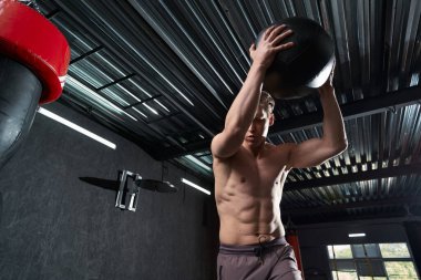 Ciddi odaklanmış zinde genç bir adam ilaç topuyla üstten vuruş egzersizi yapıyor.