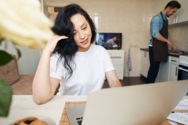 Masada oturmuş gülümseyen mutlu bayan mutfakta yemek pişirirken dizüstü bilgisayarına bakıyor.