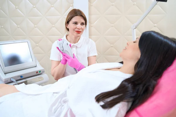 Smiling doctor with an ultrasound lifting device communicates with a female patient who is preparing for the procedure