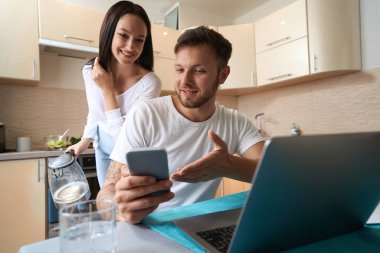 Female coming to husband from behind with kettle in hand and looking at smartphone while man showing her something