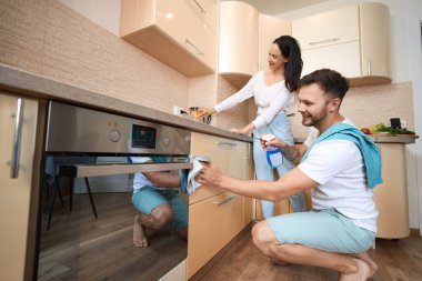 Smiling male wiping kitchen cabinet drawers with cloth and spray cleaning fluid while woman using sponge on kettle