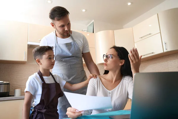 Boy and father coming to support stressed out mother during work with documents at table in kitchen