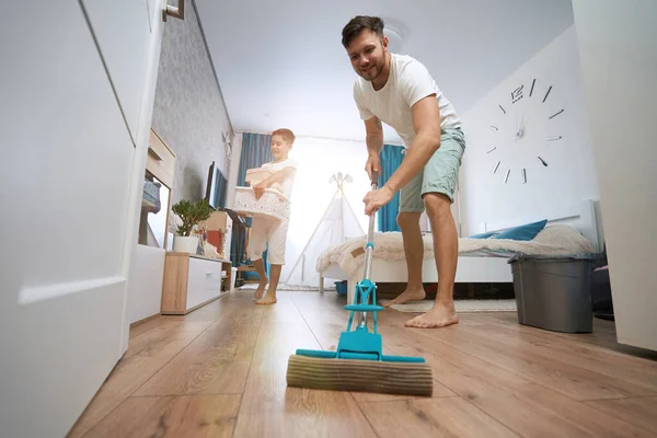 Kid bringing boxes in his hands through bedroom while man using mop for washin floor from dirt