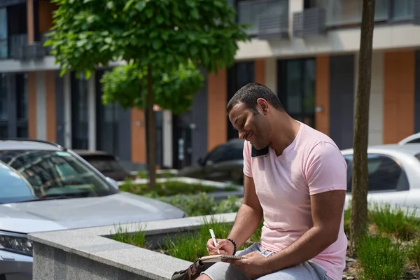 Smiling Contented Young Man Smartphone Pressed Ear Writing His Notebook — Photo