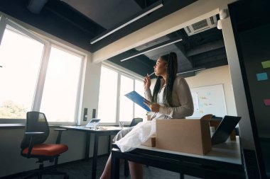 Pensive online store worker sitting on table with pen and clipboard in hands