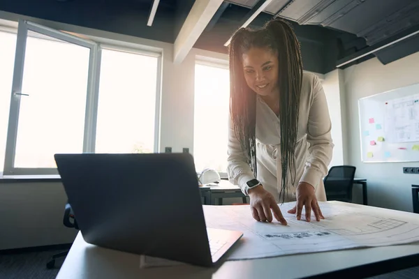 Residential design professional staring at laptop screen while leaning over rolled-out technical drawing on office desk