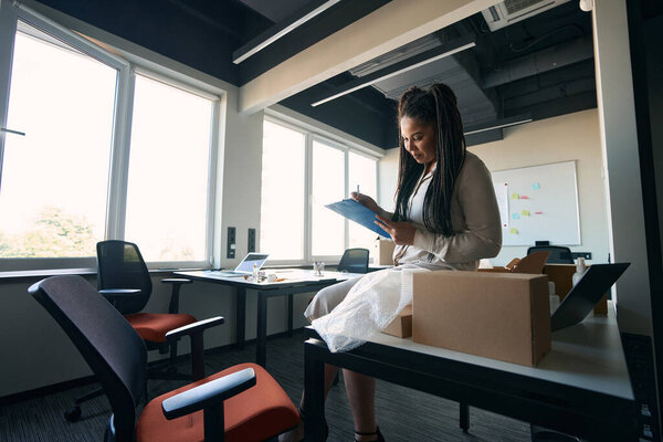 Concentrated Internet shop worker sitting on table with cardboard boxes and writing on clipboard