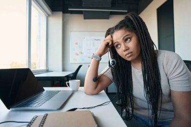 Tired female office worker with headset in hand leaning on desk and staring into distance