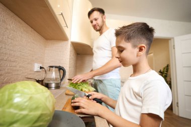 Focused Caucasian boy slicing cucumber with knife on cutting board next to his father